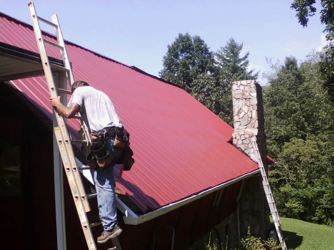 Roofer on a ladder beside a red metal roof and stone chimney — Soffit & Fascia Repair by Roofing Brothers in Princeville